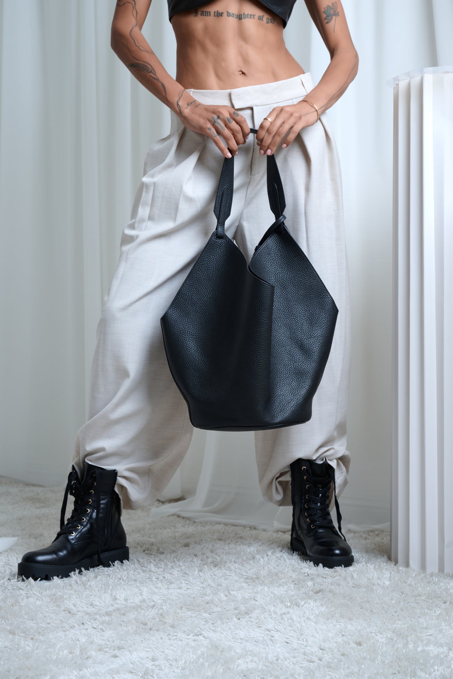Person holding a black handbag wearing white pants and black boots in a minimalistic setting.