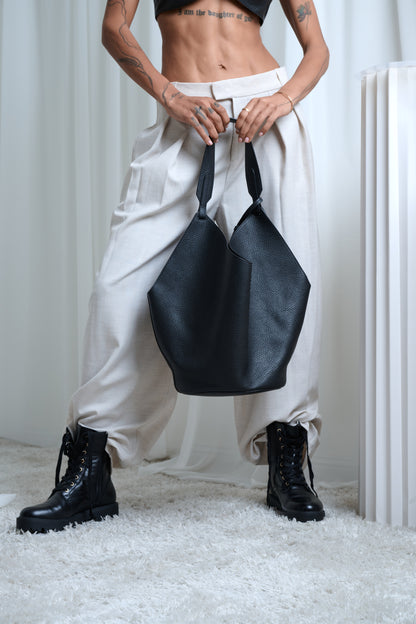 Person holding a black handbag wearing white pants and black boots in a minimalistic setting.