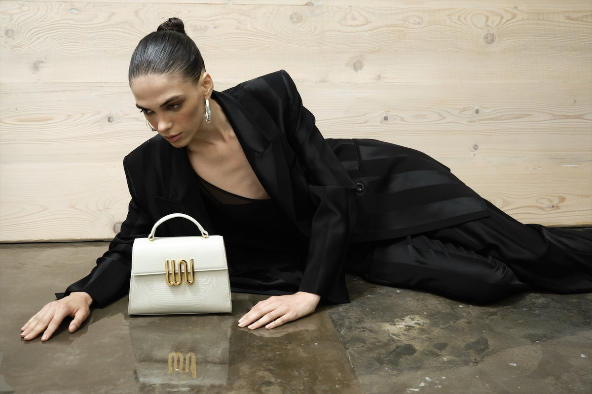 Sprawled in black tailoring on wet concrete, a pale top-handle beside her, its WN plaque reflected below.